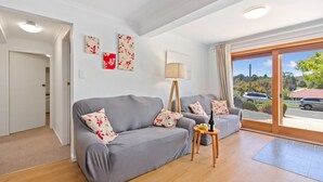 Un salón moderno con sofás grises, cojines estampados, una lámpara de pie y vista a un estacionamiento a través de amplias ventanas.