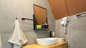 Un baño moderno con tocador de madera, lavamanos blanco, una toalla colgada en un gancho, un espejo sobre el tocador y un secador de pelo en la pared.
