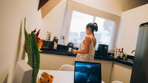 Una cocina moderna con una computadora portátil sobre una mesa, una persona cocinando y un ventanal con luz natural.
