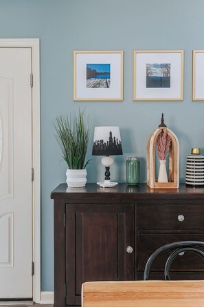 Een donkerhouten dressoir met een witte vaas, een groene vaas en een lamp met een stadslandschap silhouet. Boven de muur hangen drie ingekaderde foto's.