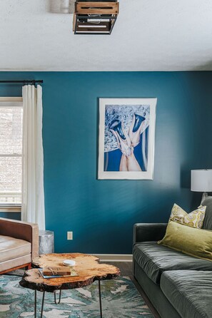A room with teal walls, a framed abstract painting, a wooden coffee table, a sofa, and a lamp.