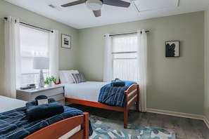 Un dormitorio con dos camas, un escritorio, un ventilador de techo y dos ventanas con cortinas.