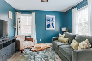 A living room with a blue wall, a grey sofa, a beige armchair, a wooden coffee table, and a TV on a cabinet. - Tucked Inn: Upper Unit - 151 Getaway/Wine + Brews (Nellysford)