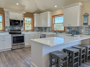 A kitchen with white cabinets, a central island with stools, a built-in oven, and a backsplash with blue tiles. - Osprey's Catch - Spacious Oceanside 8 BR Duck home w/pool, hot tub &  GOLF CART (Duck)