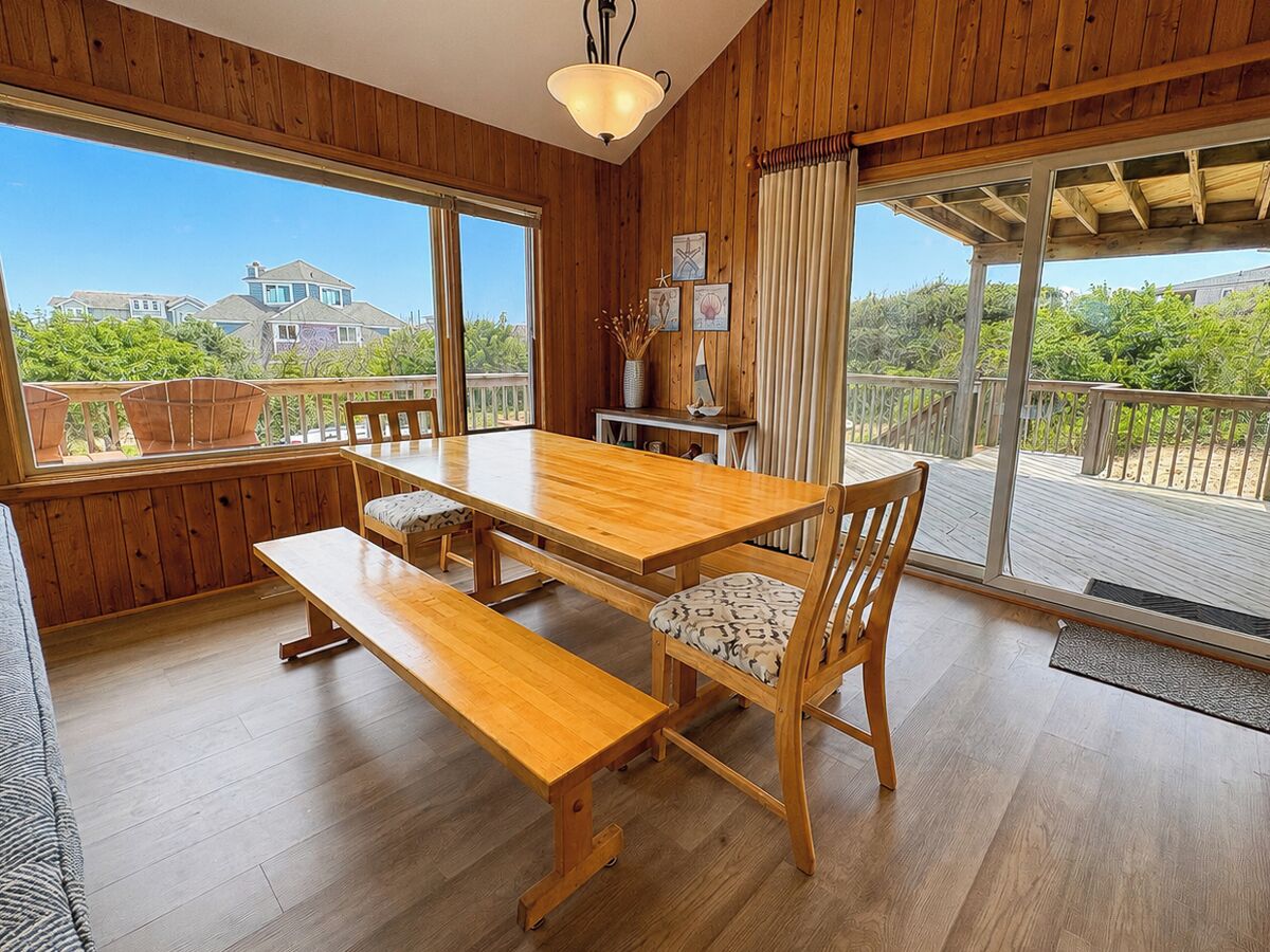 Un coin repas en bois, avec une table et des bancs, de grandes fenêtres et une vue sur la verdure et les bâtiments environnants.