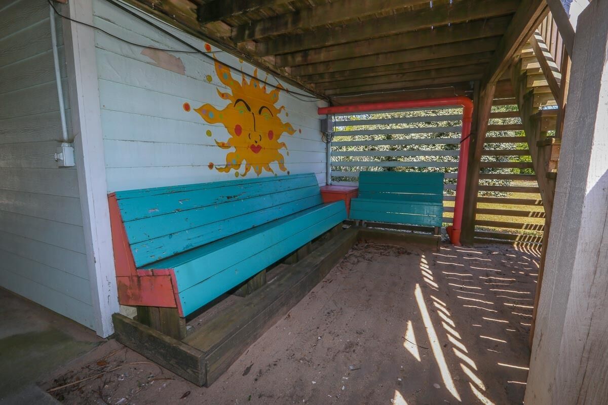 Et overdækket udendørs opholdsområde med et muralmaleri af en smilende sol på væggen, træbænke og et træhegn.