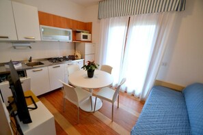 A compact room with a kitchenette, a dining table with chairs, a bed, and a window with curtains.