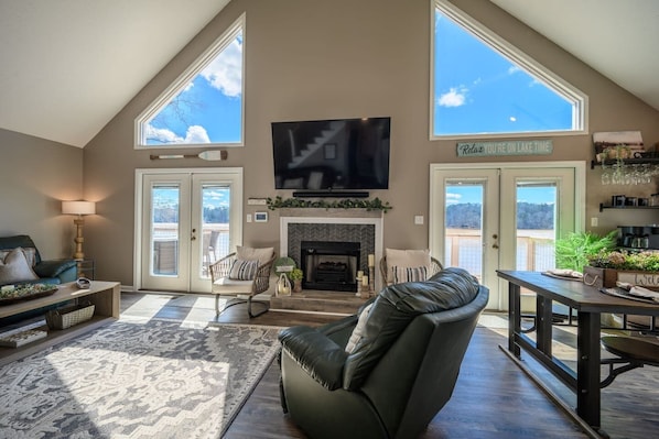 A spacious living room with a fireplace, a television mounted on the wall, and a large window offering a view of the outdoors. - Lakefront Home | Dock | Fire Pit | Pool Table (Eatonton)