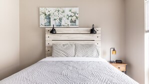 Un dormitorio con una cama, cabecera con un cuadro, lámparas de noche y mesita de noche con un reloj.