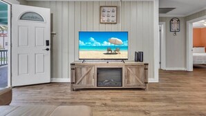 A living room with a large flat-screen TV on a wooden console, a door with a decorative glass panel, and a framed picture above the TV. - Amazing Home on Main St - Walk to the Beach (North Myrtle Beach)