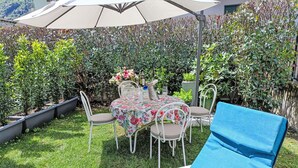 A patio with a table and chairs, surrounded by lush greenery and a large umbrella. - Olmo Garden Apartment by Interhome (Valtellina)