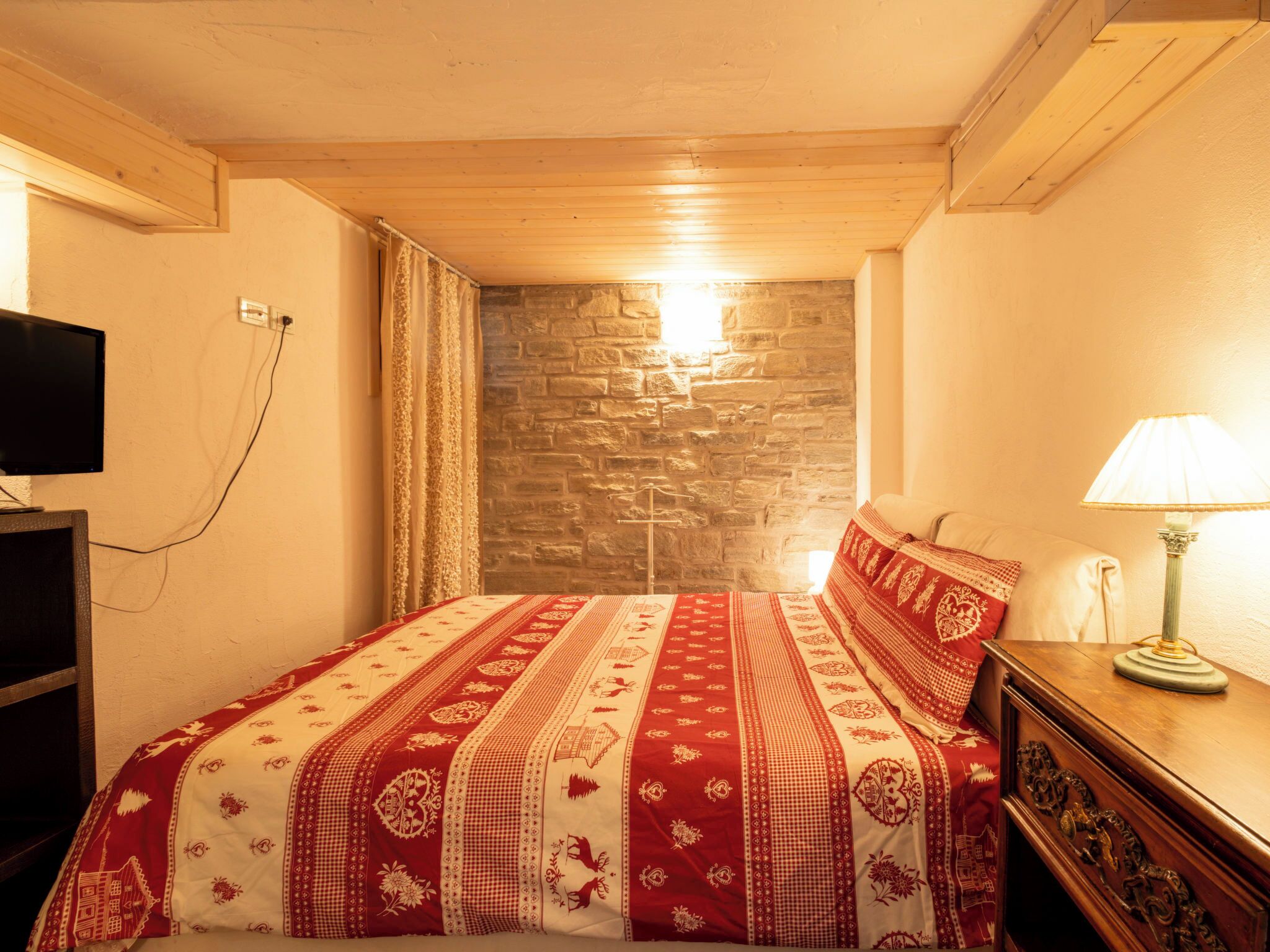 A bedroom with a stone wall, a wooden ceiling, a bed with a patterned bedspread, a TV, and a lamp.
