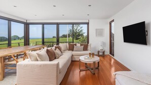 Un salon spacieux avec un grand canapé en forme de L, une table à manger en bois et une table basse ronde. Une grande fenêtre offre une vue sur un paysage verdoyant.