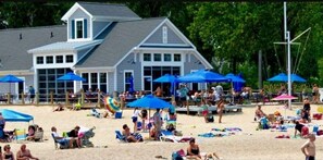 Un edificio frente a la playa con varios sombrillas azules, sillas de playa y gente relajándose.
