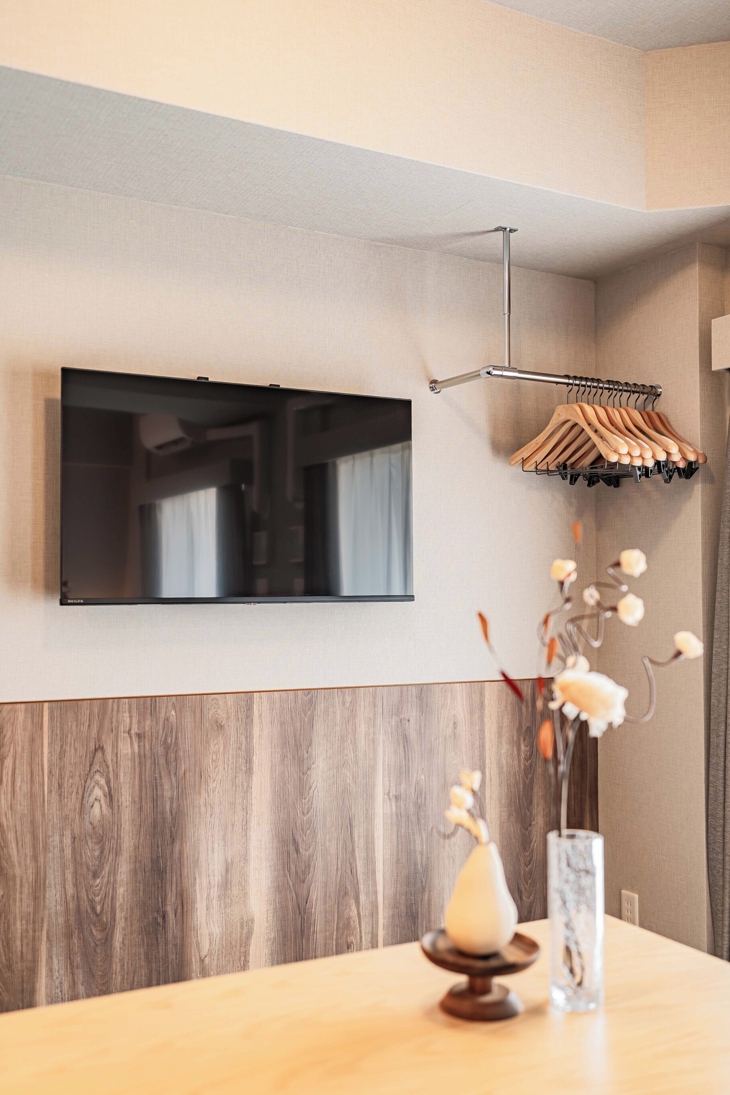 Ein modernes Zimmer mit einem Wand-Fernseher, einem Holztisch mit Vase und Kerze sowie einem Kleiderständer mit Kleiderbügeln.