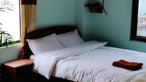 Une chambre à coucher comprenant un lit, une table de chevet, une fenêtre avec des rideaux, une étagère fixée au mur et une vue sur les bâtiments à l’extérieur.