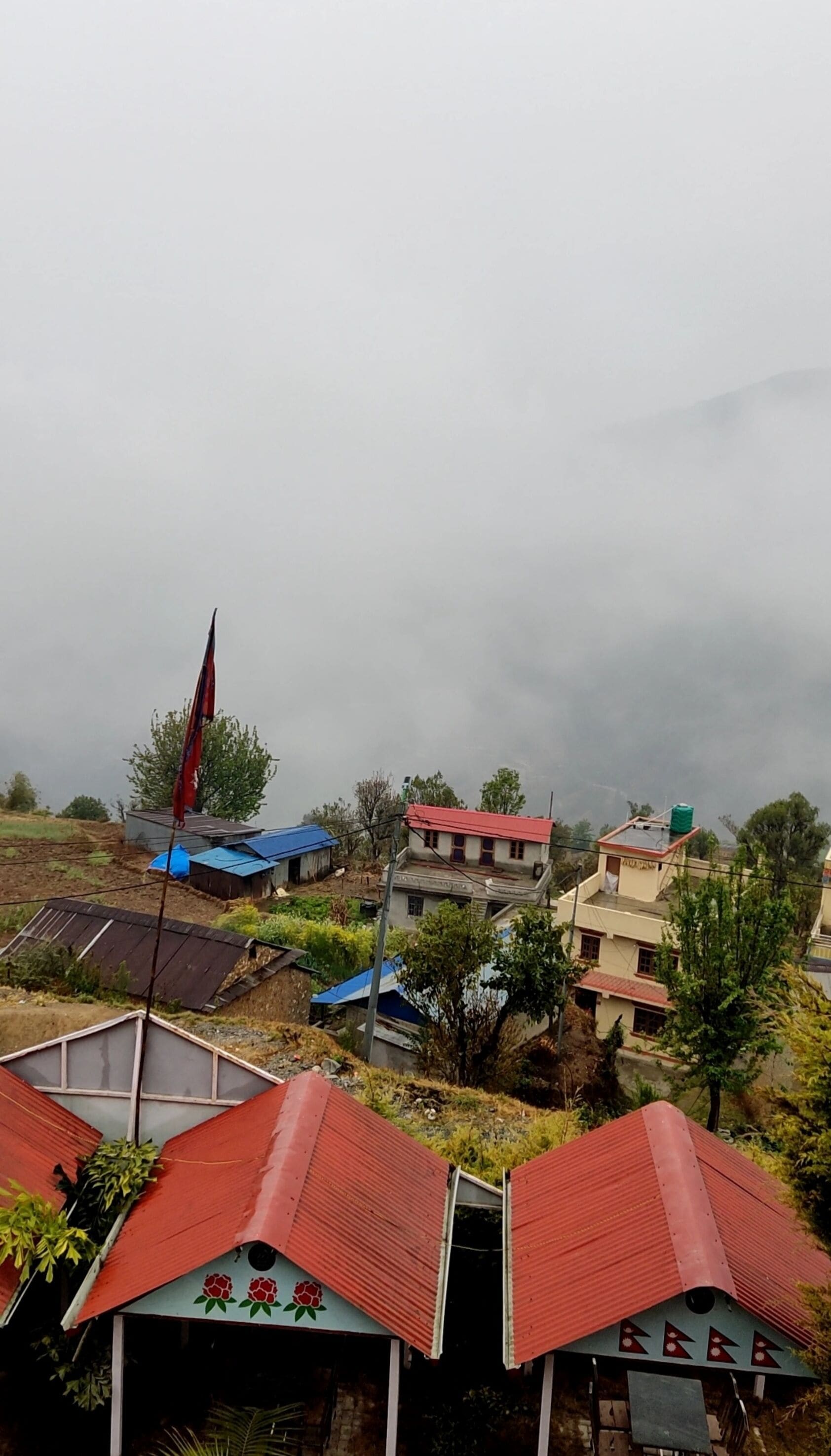 Un pueblo rural con casas de techos rojos, una bandera en un asta y montañas al fondo.