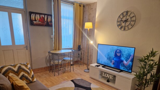 A living room with a sofa, a TV on a stand, a dining table with chairs, a lamp, and a clock on the wall. - House - Sleeps 4 - Garden - Parking (Wallsend)