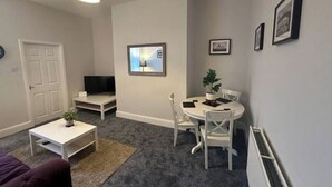 Un espacio habitable compacto con una puerta blanca, un televisor de pantalla plana, una pequeña mesa blanca, una mesa de comedor con sillas y una planta en maceta.