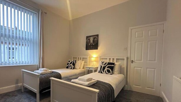 Un dormitorio con dos camas individuales, una mesita de noche con lámpara, una puerta y una ventana con persianas.