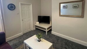 Una sala de estar con una mesa de centro blanca, un televisor de pantalla plana sobre un mueble, una puerta y un cuadro enmarcado en la pared.