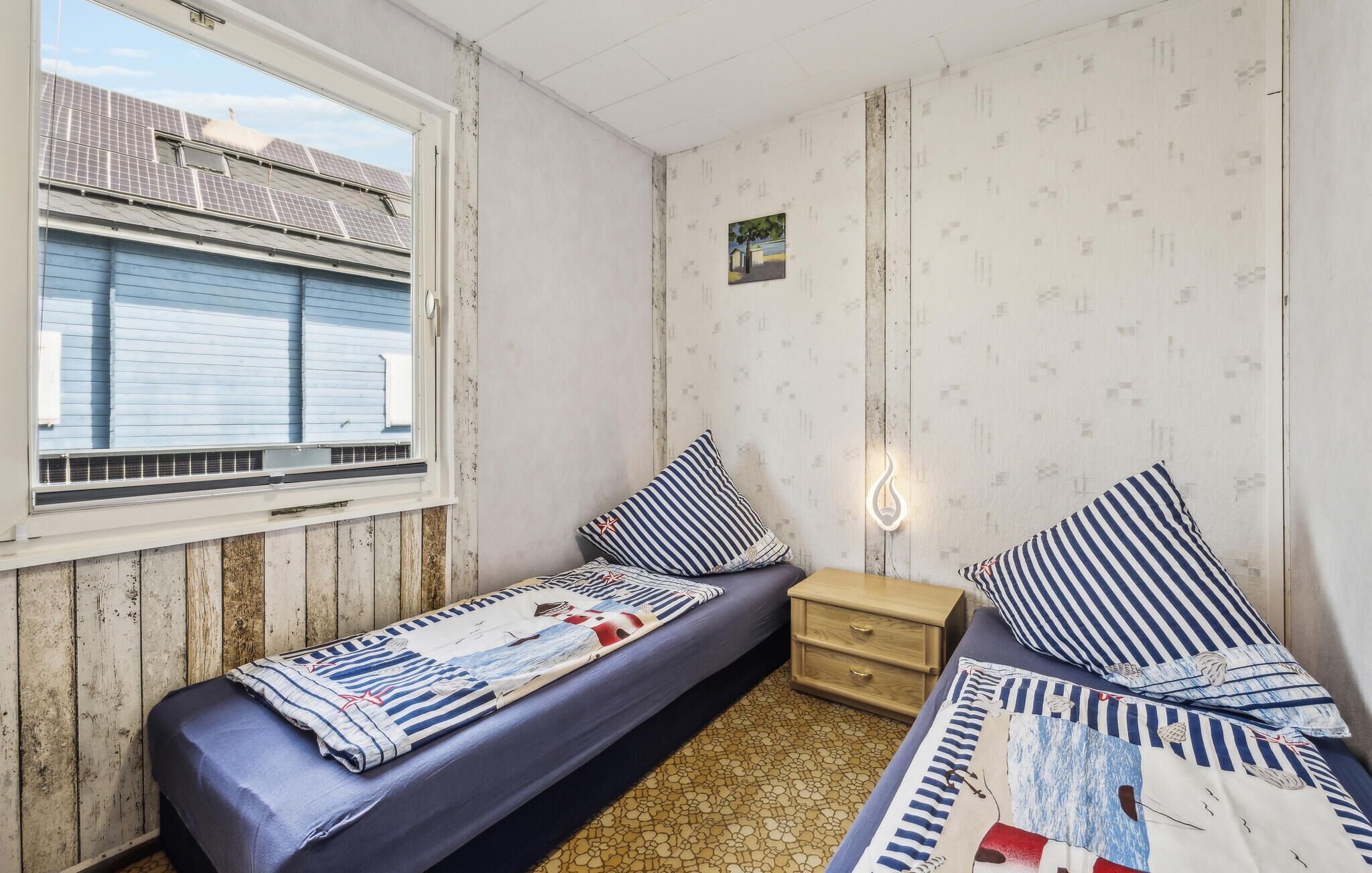 A small, single-bed room with a window, wooden walls, a small bedside table, and striped bedding.