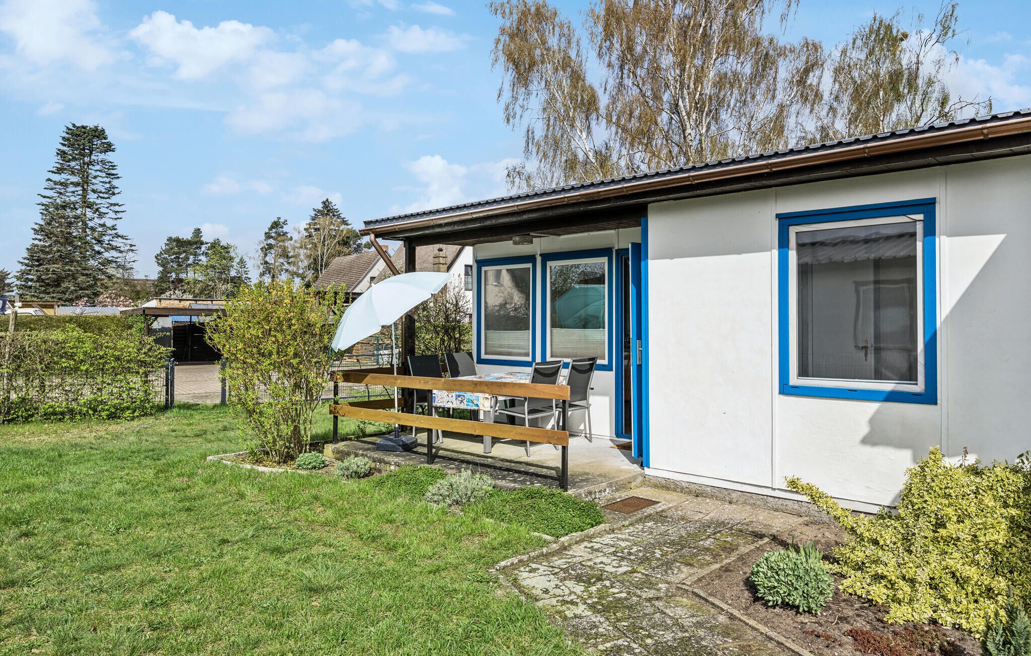 Une petite maison à un seul étage, avec une fenêtre à cadre bleu et une porte, un banc en bois à l’extérieur et un parasol blanc.