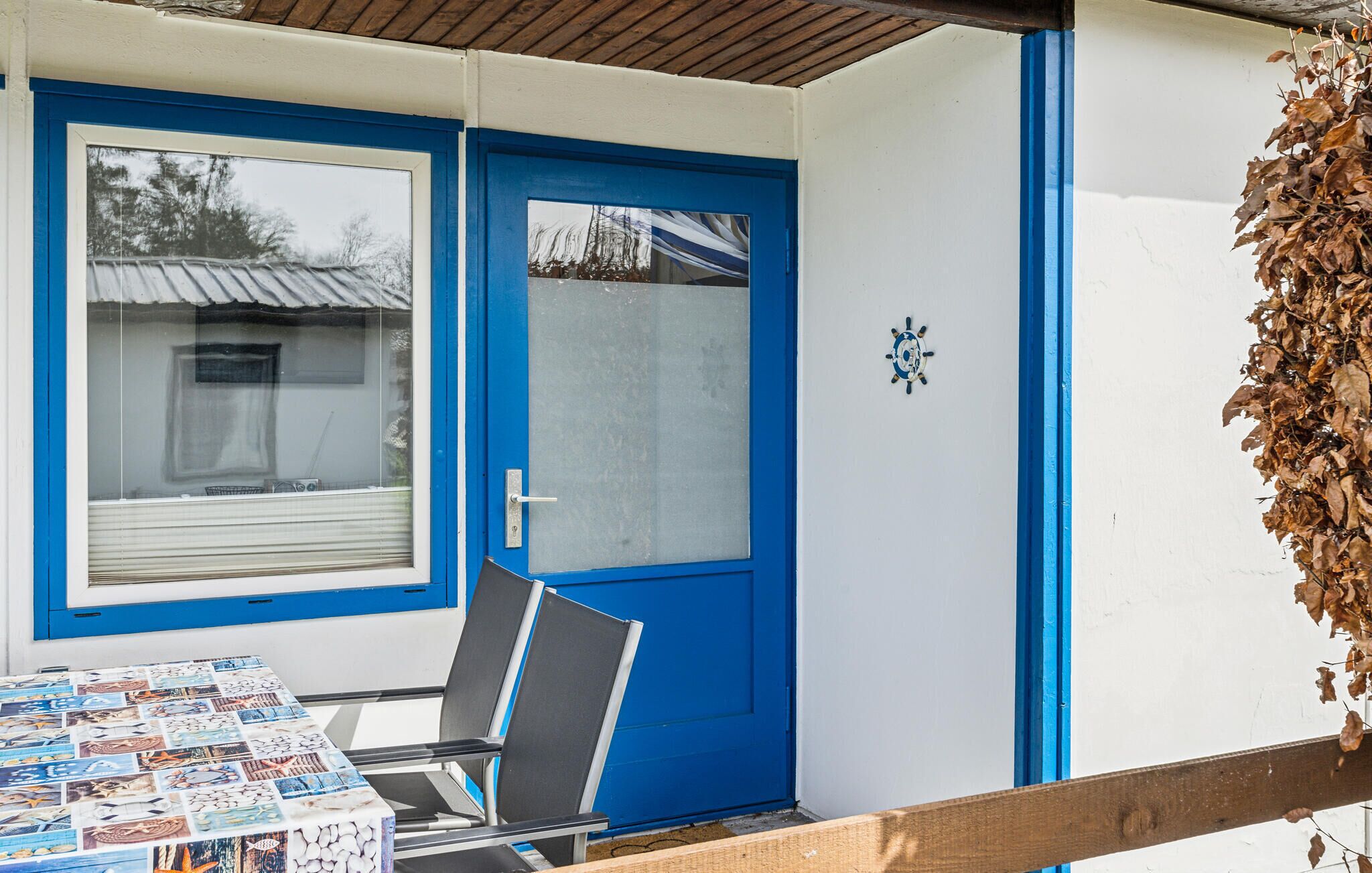 A room with a blue door, a window with a view of a building, a table with a magazine rack, and a chair.