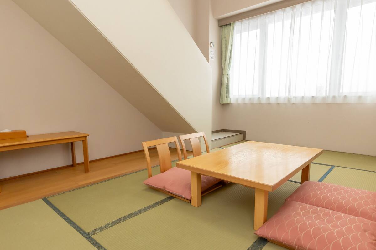 A room with tatami flooring, a low wooden table, a bench, and a window with sheer curtains.