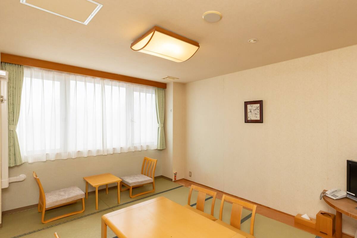 A room with a wooden table and chairs, a window with sheer curtains, a small framed picture on the wall, and a telephone on a wooden desk.