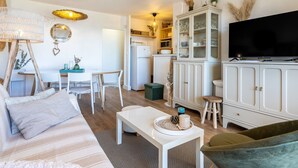 Un salon moderne avec un canapé, une table basse et un téléviseur. On y trouve également un coin repas avec des chaises et une table. Une cuisine équipée de placards et d’un réfrigérateur est visible.