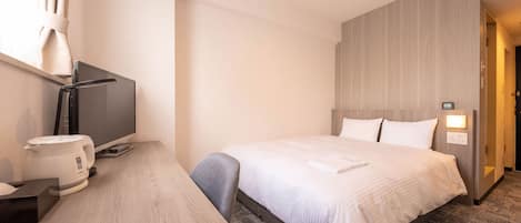 A hotel room with a bed, a desk with a computer and a coffee maker, a TV, and a wall-mounted light.
