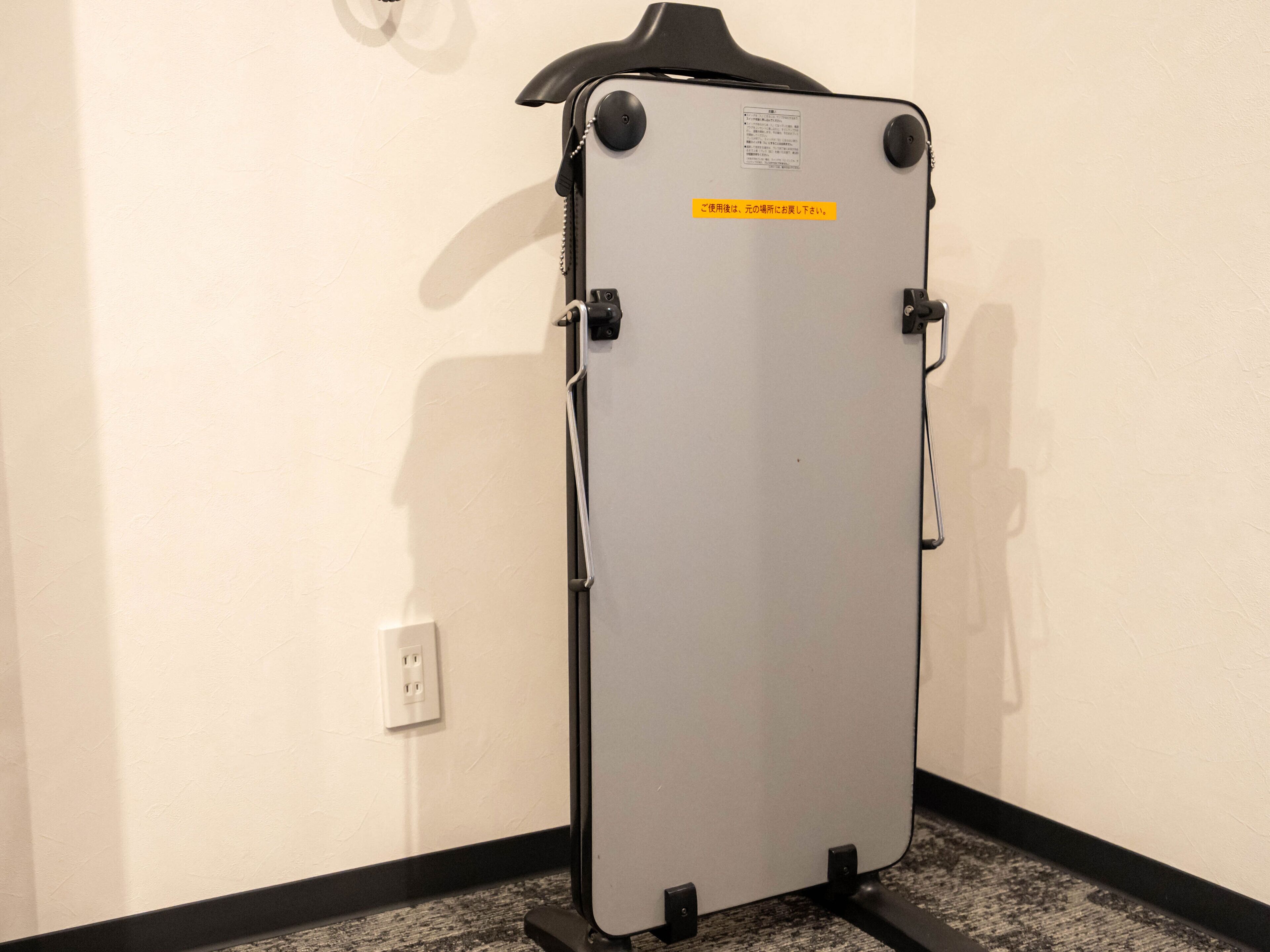 A portable, metallic, rectangular object with a black top and a yellow label on the back, mounted on a black stand against a white wall with a black baseboard.
