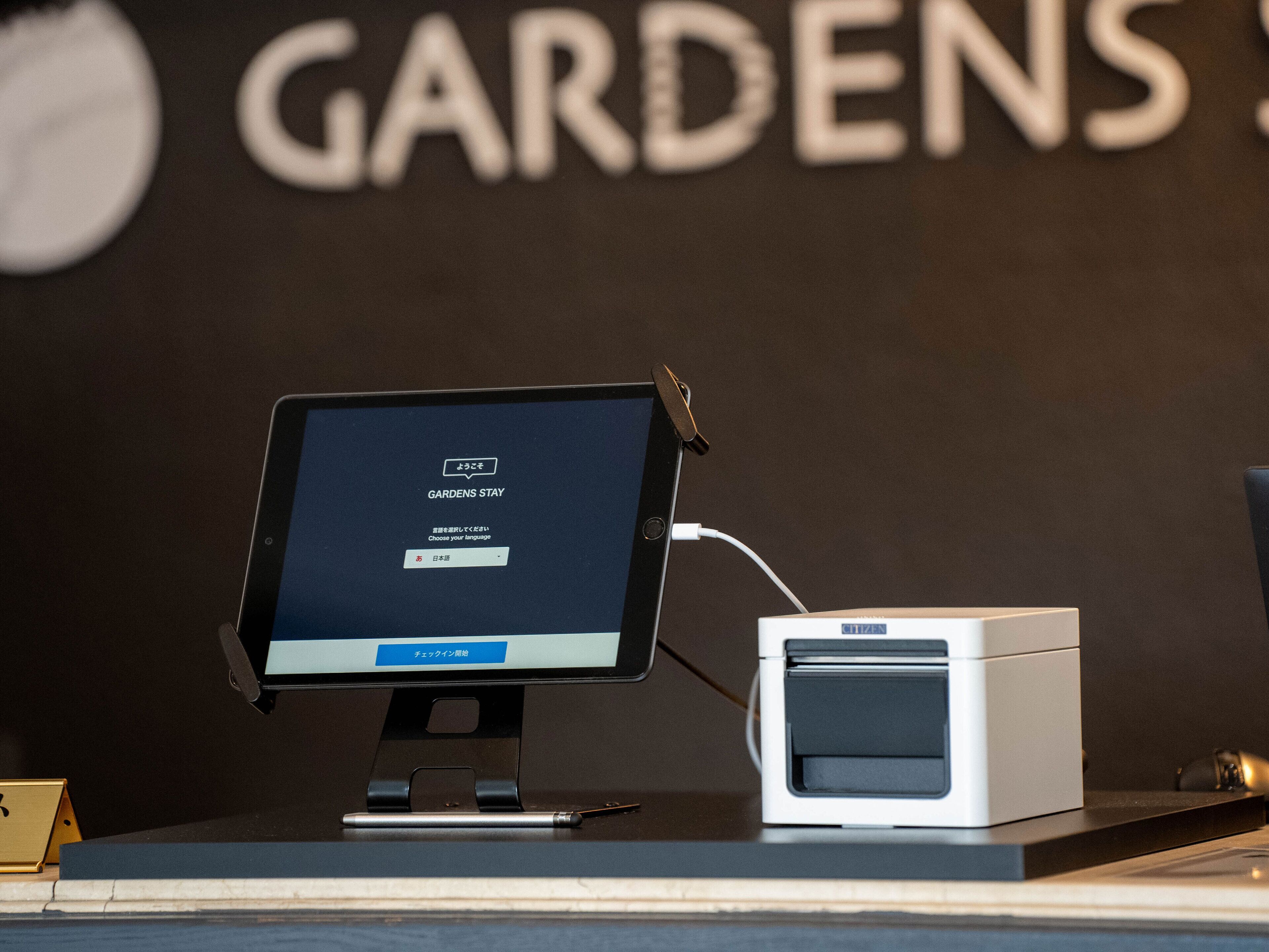 Un monitor de computadora sobre un soporte, con teclado y ratón, una impresora, y una pared donde se lee la palabra "GARDENS".