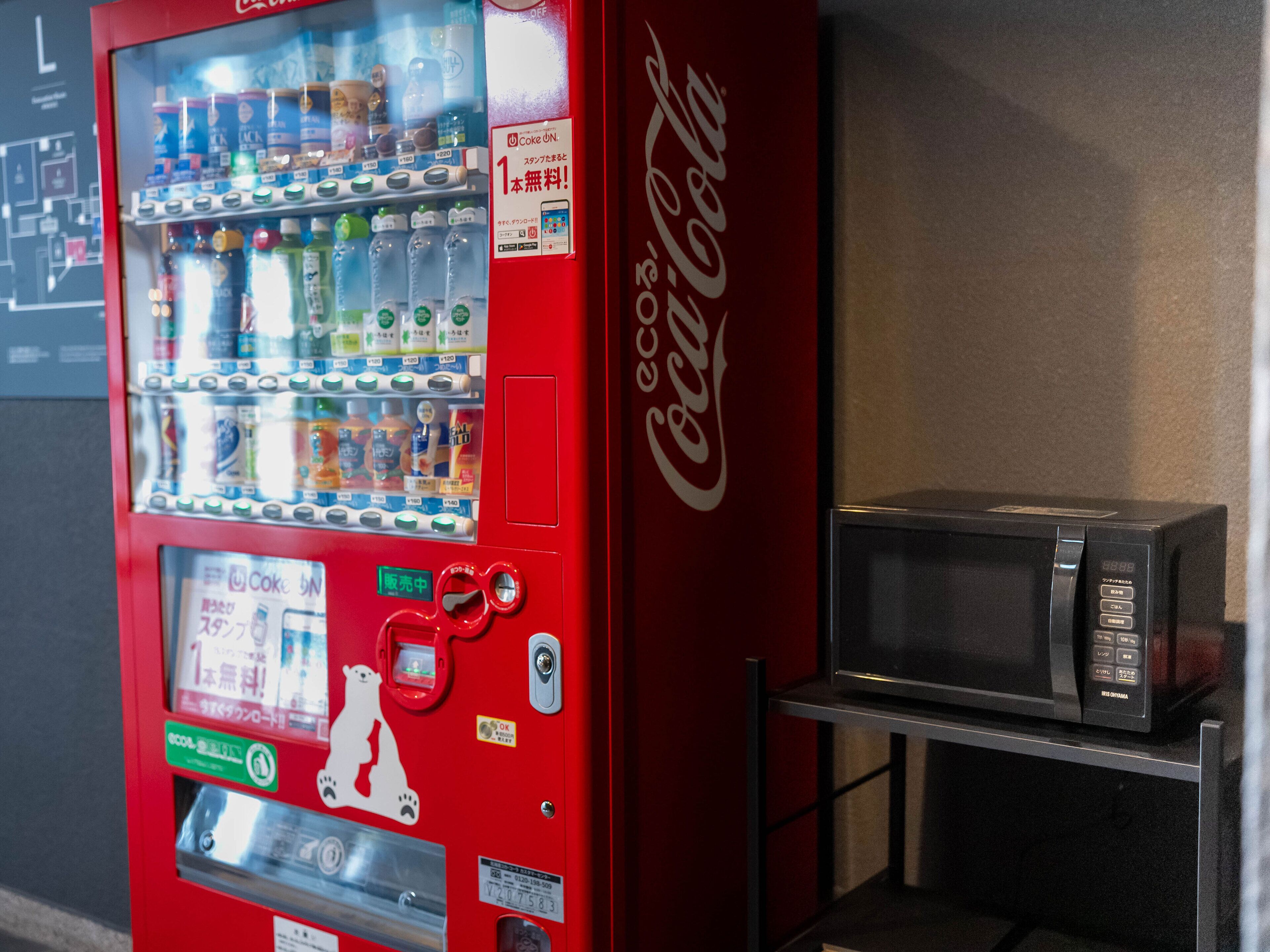 A vending machine with a Coca-Cola logo, a polar bear sticker, and a microwave on a shelf.