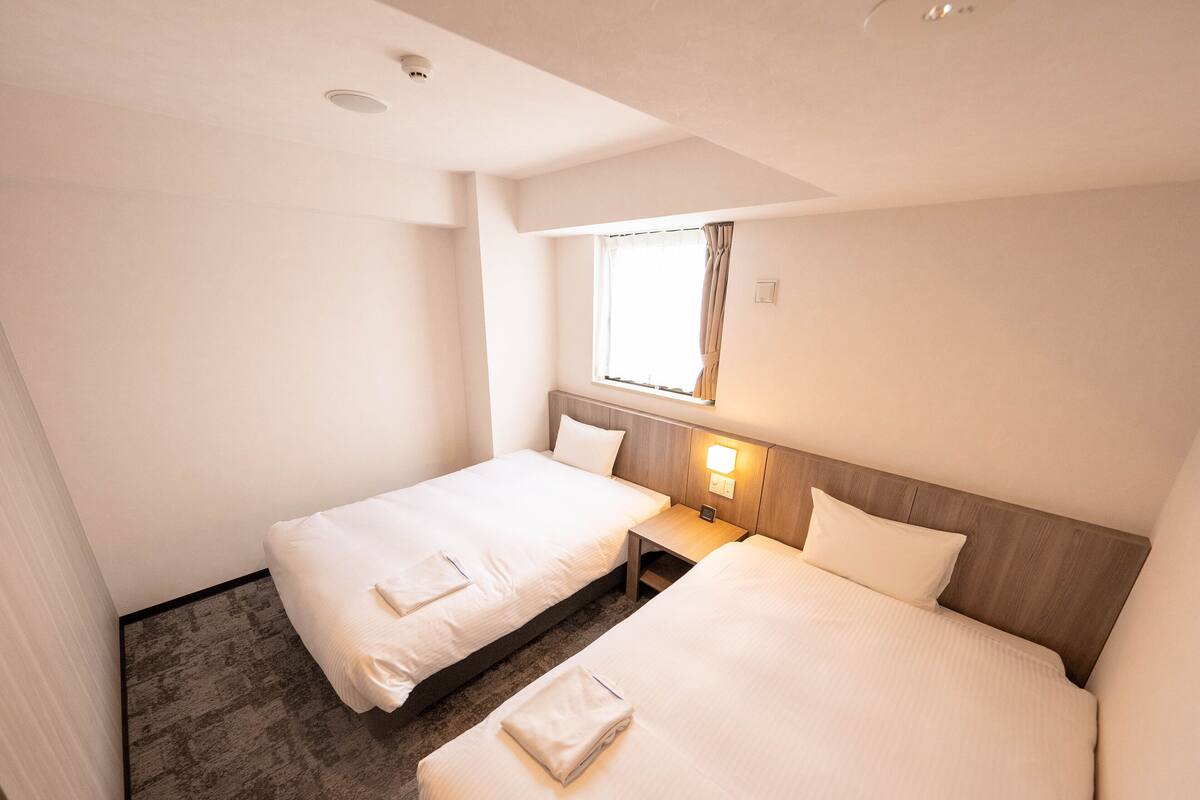 Une chambre d’hôtel avec deux lits simples, une petite table de chevet avec une lampe, une fenêtre avec des rideaux et un luminaire au plafond.