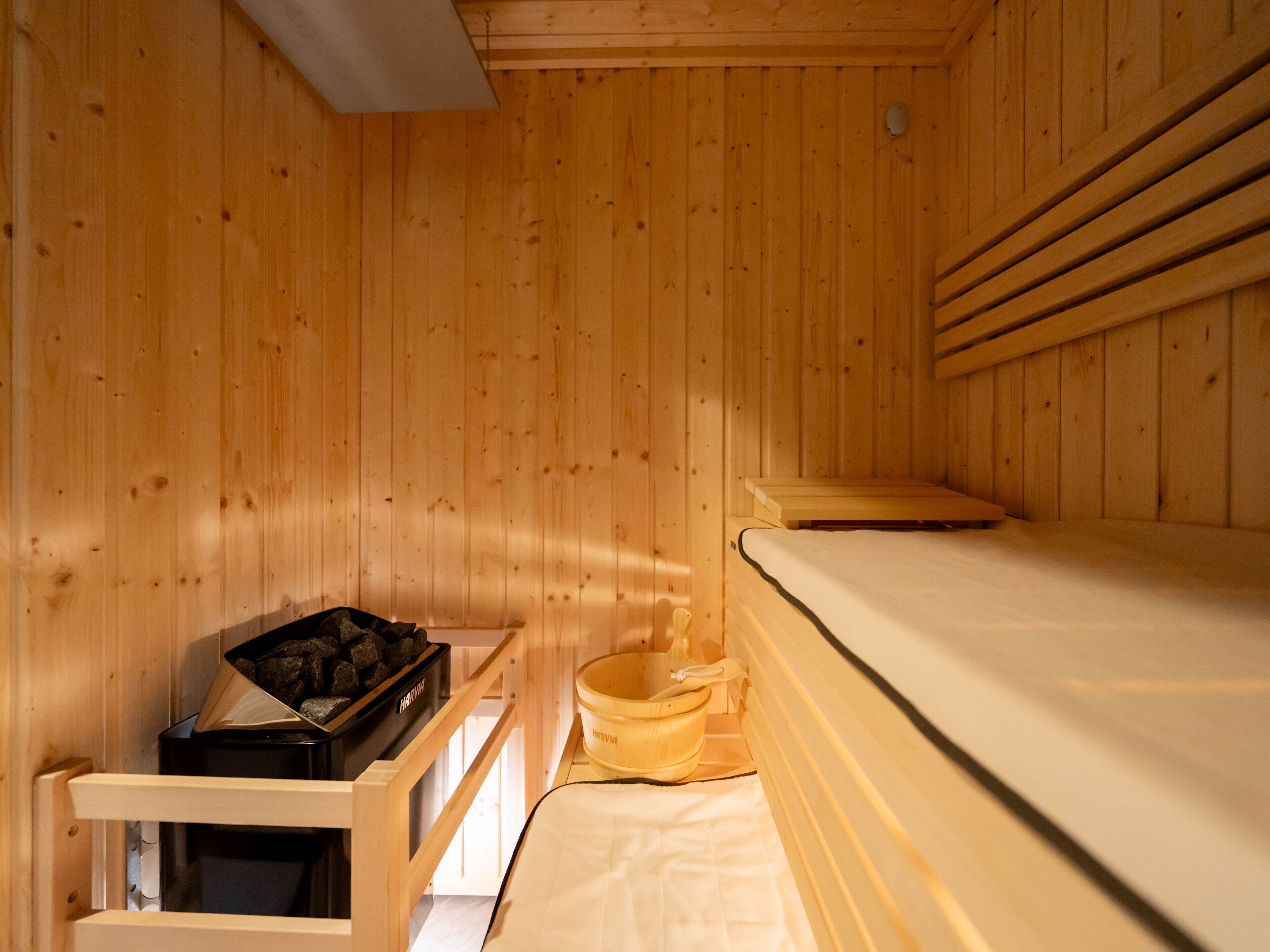 A wooden sauna room with a bed, a wooden bench, and a small table with a trash bin.