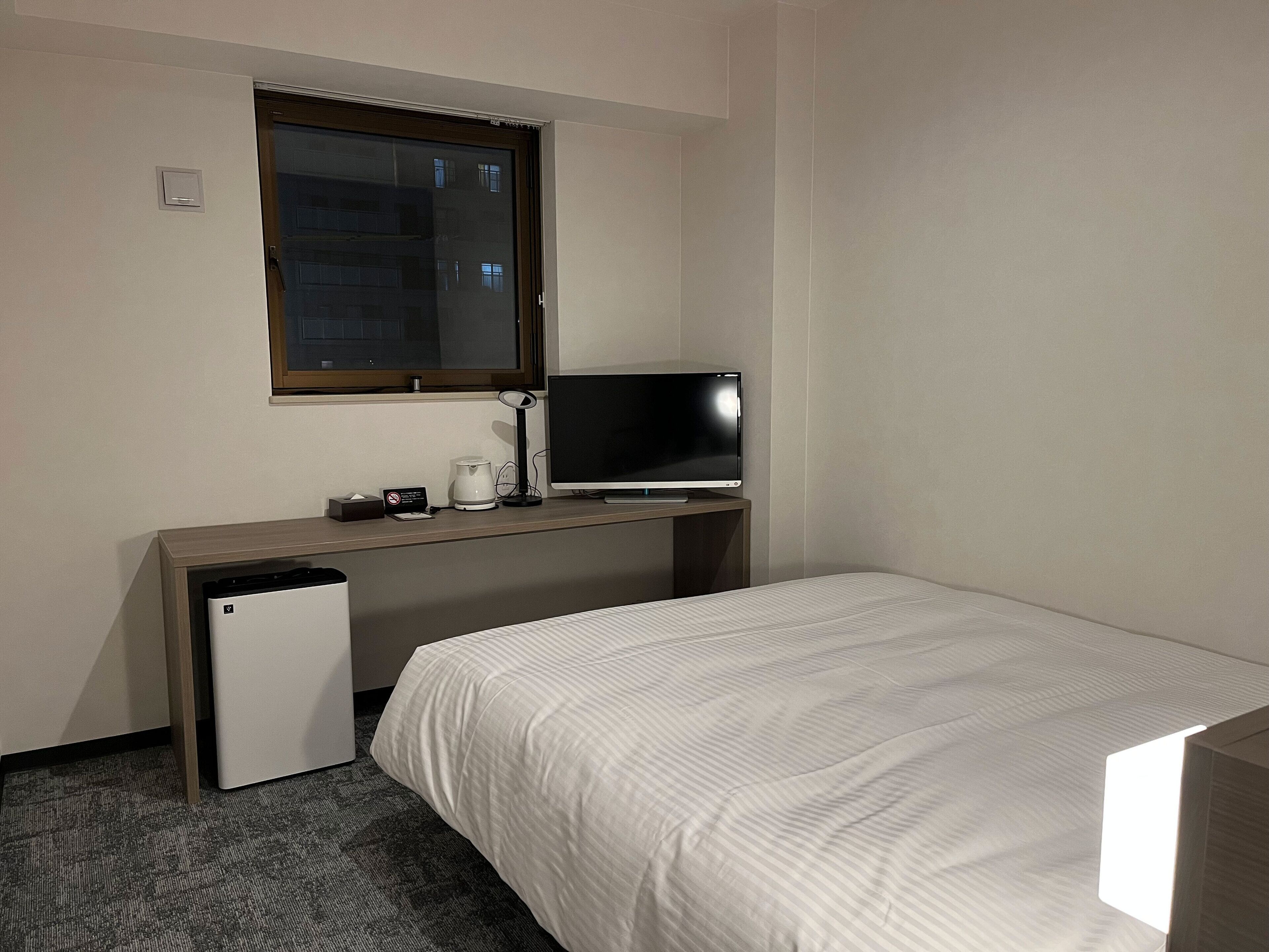 Ett hotellrum med en säng, ett skrivbord med en datorskärm, en kaffebryggare och ett litet kylskåp.