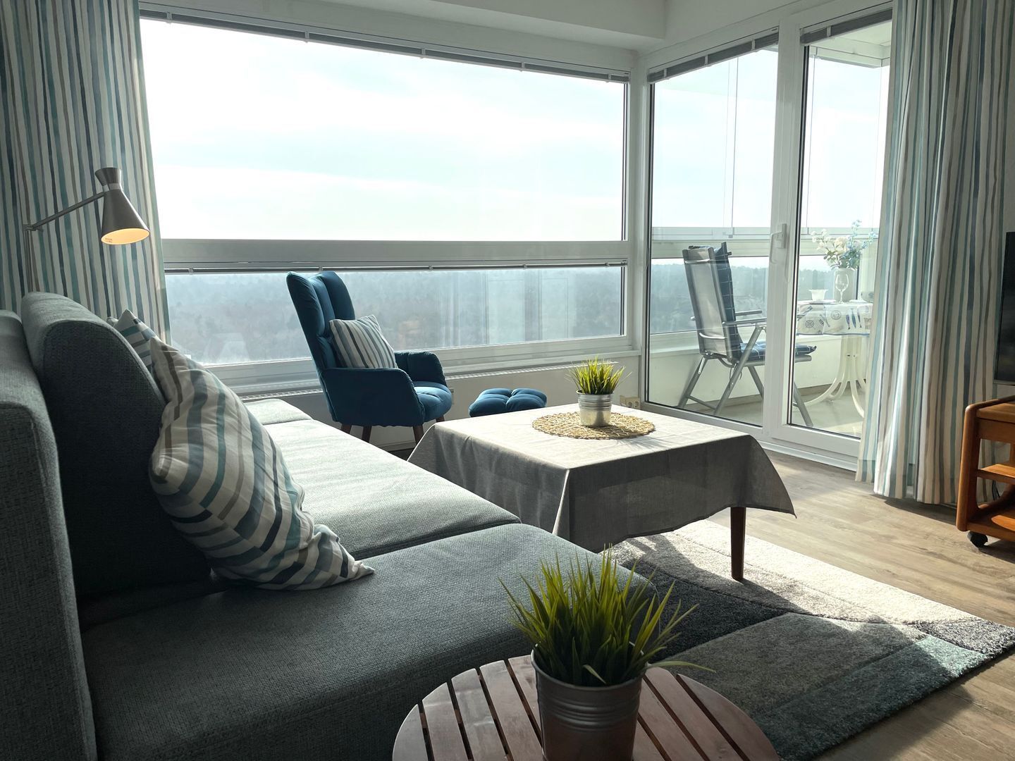 A modern living room with a grey sofa, striped pillows, a blue armchair, a small table with a plant, and a view of the balcony.