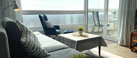 A modern living room with a grey sofa, striped pillows, a blue armchair, a small table with a plant, and a view of the balcony.