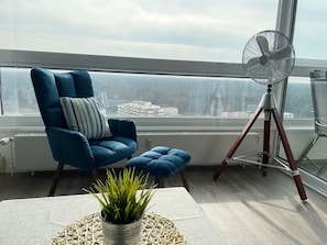 A modern balcony with a blue chair, a small table with a potted plant, and a standing fan.