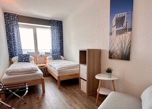 A hotel room with two single beds, a small table, a chair, a wardrobe, and a window with curtains.