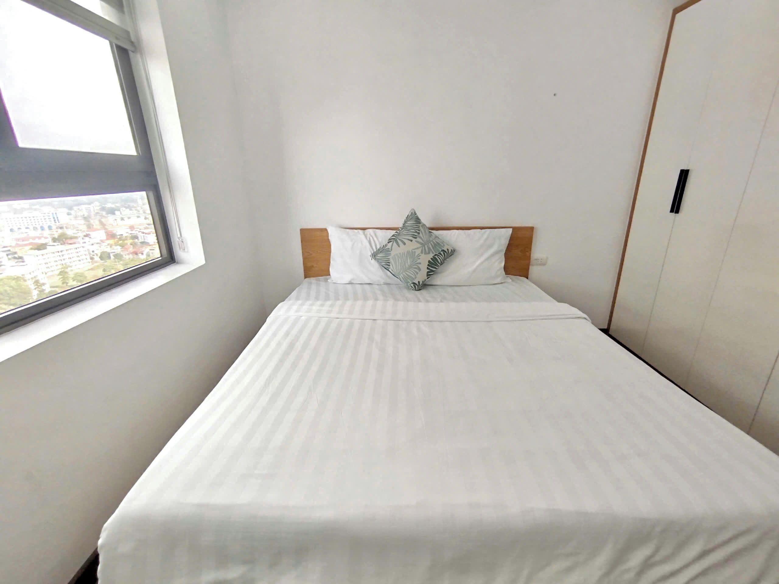 A single bed with a white bedspread and a leaf-patterned pillow, a wooden headboard, and a window with a view of buildings.