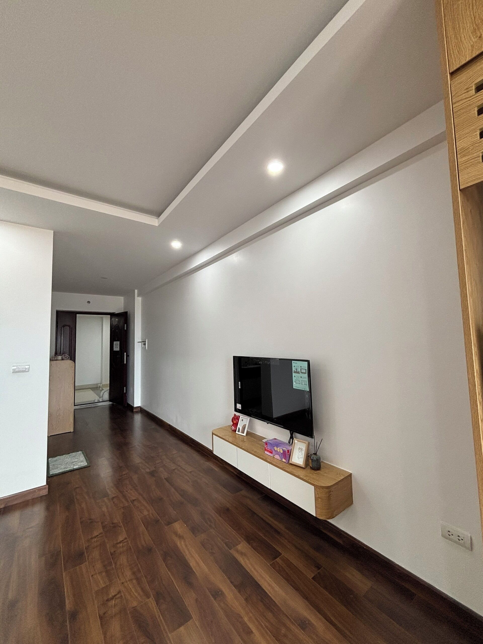 A modern living room with a flat-screen TV on a wooden console, a wooden floor, and a white ceiling with recessed lighting.