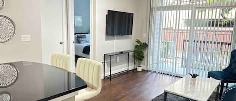 A modern living room with a flat-screen TV, a dining area with a glass-top table, and a large window with blinds.