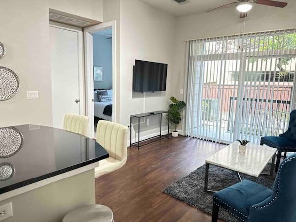 A modern living room with a flat-screen TV, a dining area with a glass-top table, and a large window with blinds.