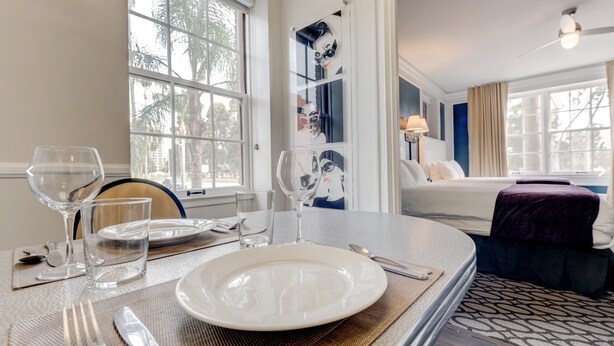 Habitación de hotel con comedor listo para dos personas, cama con cabecera azul y ventana grande con vista a palmeras.