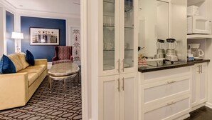 Una sala moderna con un sofá amarillo, alfombra estampada y una silla roja. Junto a ella, una cocina con armarios blancos, encimera negra y una cafetera.