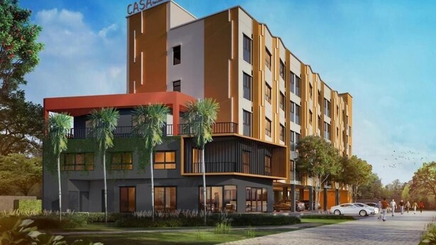 A modern multi-story hotel with a prominent red and beige facade, multiple balconies, and a landscaped front yard.