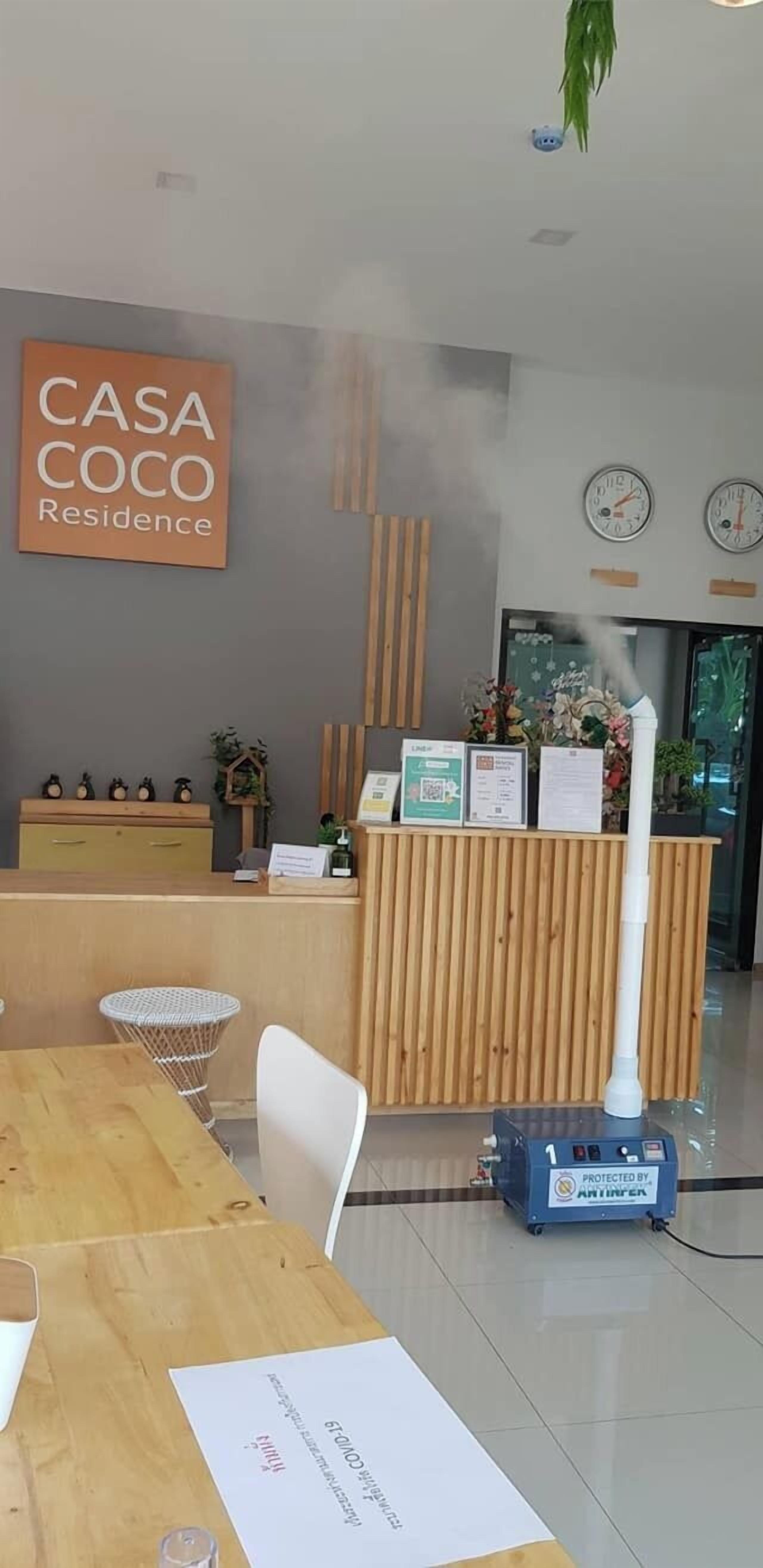Vastaanottoalue, jossa on puinen työpöytä, kyltti, jossa lukee ”CASA COCO Residence”, ja höyrypesuri.